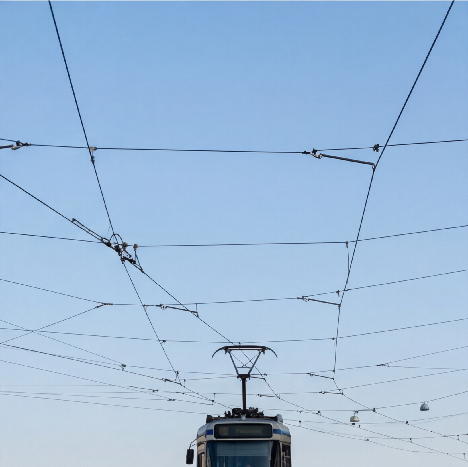 Husté trolejové vedení tramvaje na modré obloze, dole vidět přední část přijíždějící tramvaje